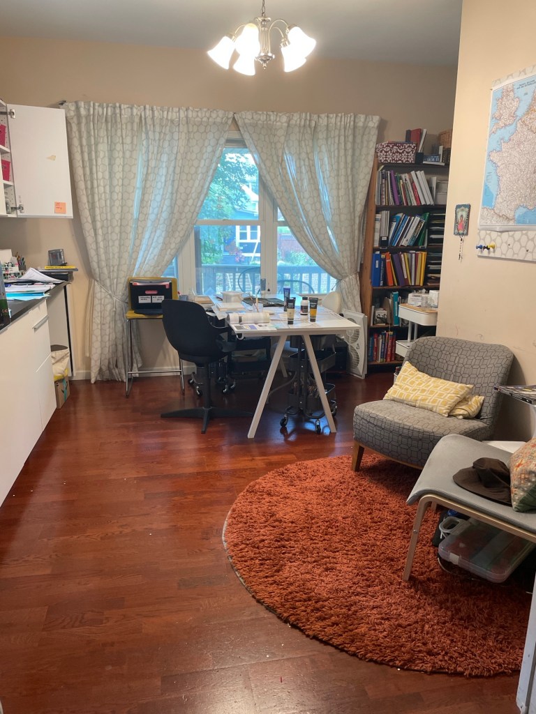 A room with a curtained window at the far end looking out on the street.  A white table with a half-finished painting project spread out.  Two comfy chairs in the foreground, on a round area rug the same color as the wood floor.  A countertop and cabinetry, a stuffed bookshelf, a map of France on the bulletin board.