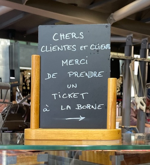 hand-chalkes sign saying “chers clientes et clients merci de prendre un ticket à la borne” with an arrow pointing 