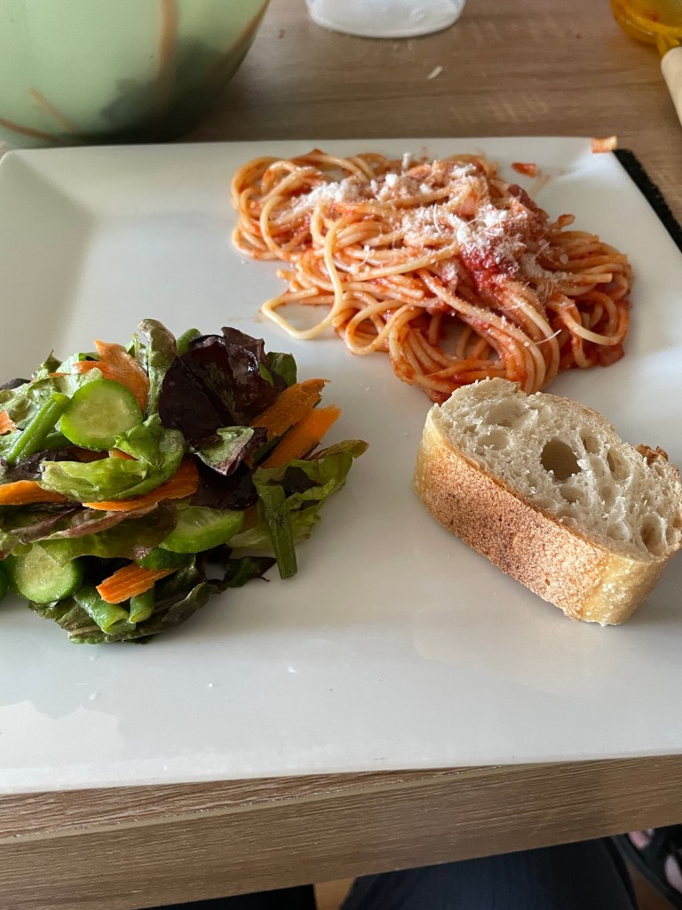 Square white dinner plate with a pile of red-sauced, Parmesaned pasta in one corner, a pile of green salad in another corner, and a chunk of baguette in another.