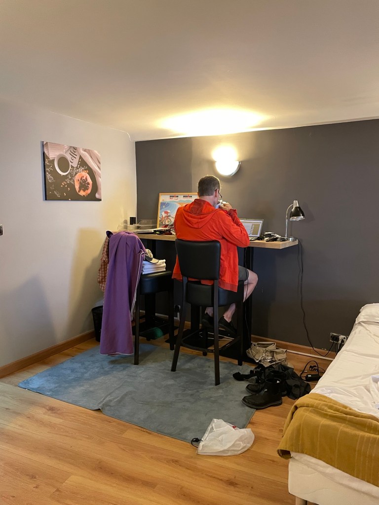 A man in a bright orange hoodie sits with his back to the camera on a high stool at a bar-height table against a wall in a bedroom.  He is working on a laptop.