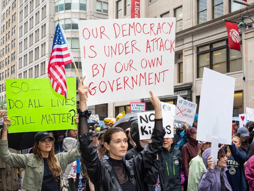 A crowd of protesters in New York City, a library in the background, holding signs.  An American flag is also being carried.  Visible signs read:  "So... do all lives still matter?"  "Our democracy is under attack by our own government" "We the People say No Kings" "No Kings" 
