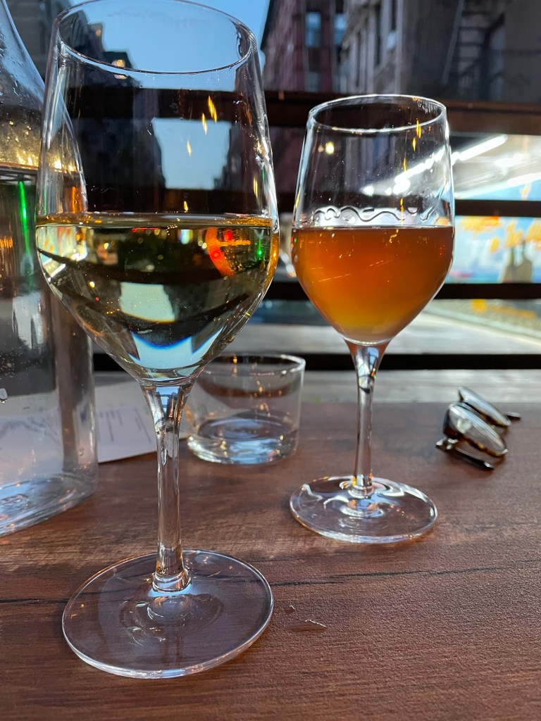 Two glasses of wine on a table, on an outdoor restaurant table, with the glow of sky at dusk behind.  One is white wine, one is a rosy orange.  There is a pair of glasses folded on the table.