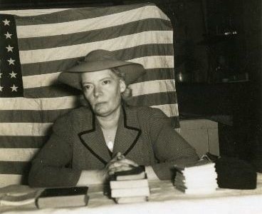Dorothy Day seated in front of a large U.S. flag and behind a table on which are a few stacks of books.  She wears a hat and a light-colored suit jacket with dark piping along the lapel.  Her forearms and clasped hands rest on the table.  Her expression is weary.